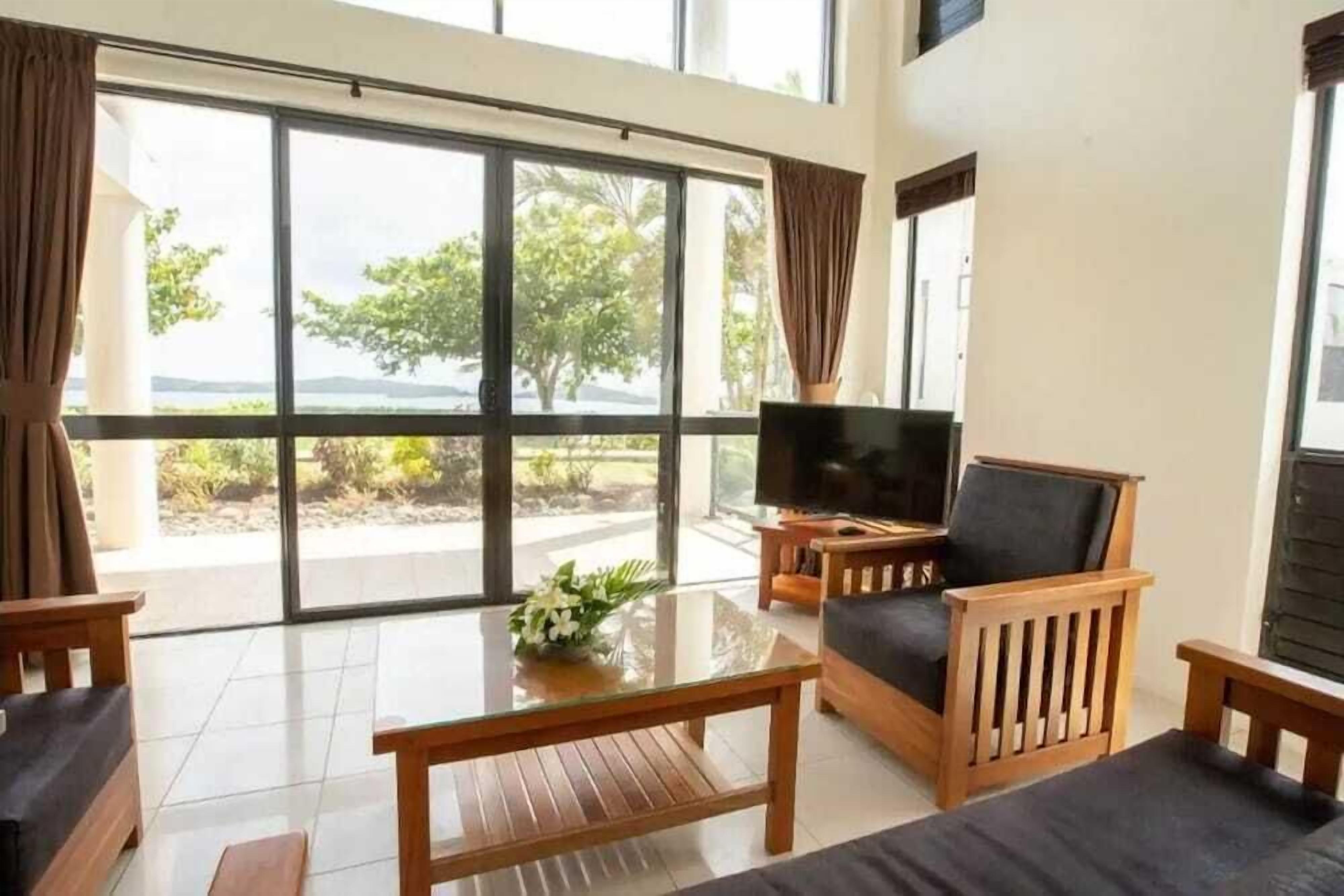 A spacious living area with a large sliding glass door leading to a balcony, a flat-screen TV, and a wooden coffee table with a vase of flowers.