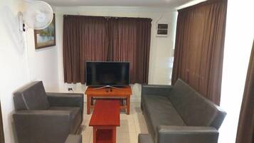 A living room with a flat-screen TV, a sofa, a coffee table, and a fan.