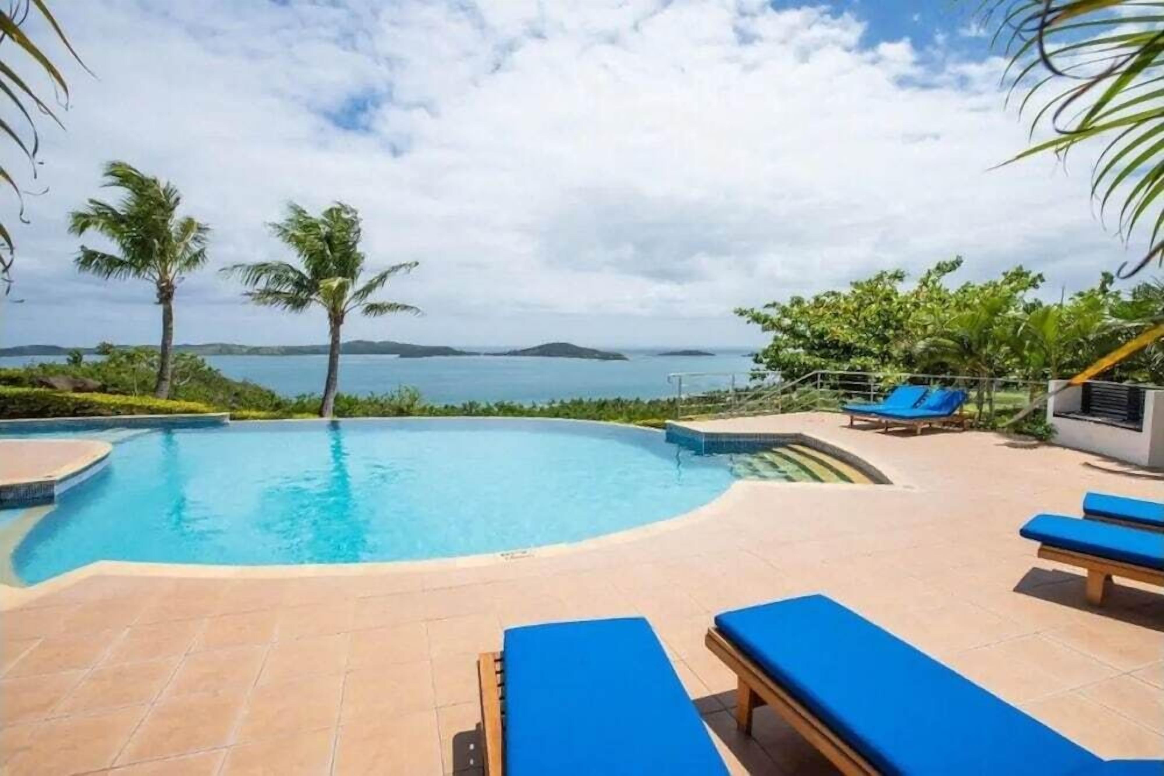 Una piscina con borde curvo, rodeada de sillones de descanso y palmeras, con vista al mar y a islas distantes.