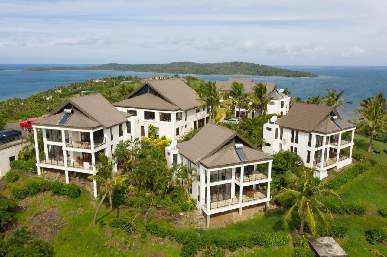 A coastal property with multiple white buildings featuring large windows and balconies, surrounded by lush greenery and a view of the ocean.