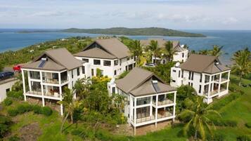 A coastal property with multiple white buildings featuring large windows and balconies, surrounded by lush greenery and a view of the ocean.