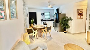 Uma sala de estar moderna com sofá, mesa de jantar com cadeiras, área de cozinha com geladeira e ventilador de teto.
