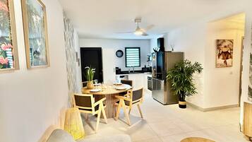 Een moderne woonkamer met een bank, een eettafel met stoelen, een keuken met een koelkast, en een plafondventilator.