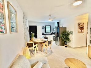 Ett modernt vardagsrum med en soffa, ett matbord med stolar, en köksdel med ett kylskåp och en takfläkt.