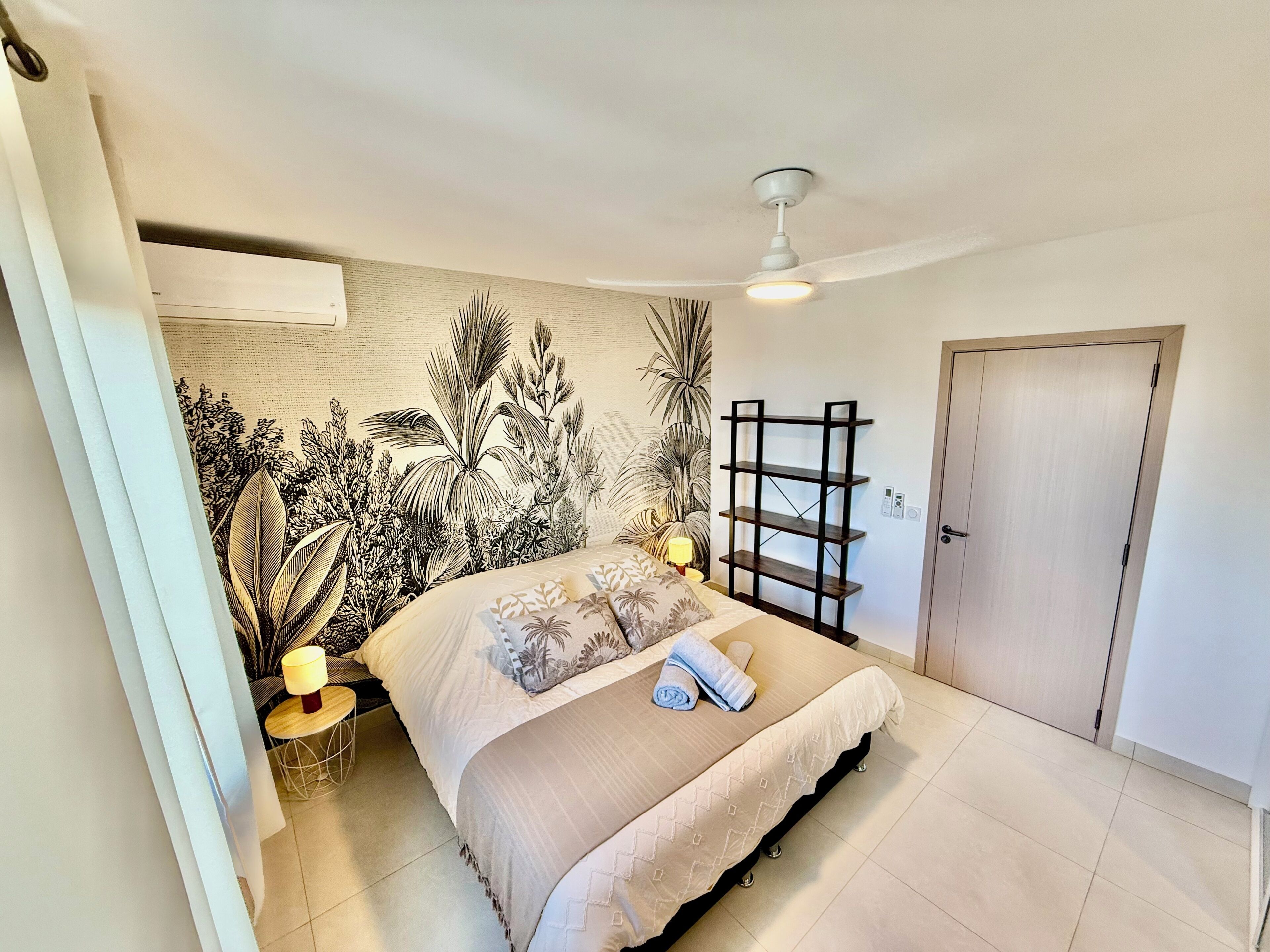 A bedroom with a bed, a nightstand, a wall-mounted shelf, a ceiling fan, and a door.