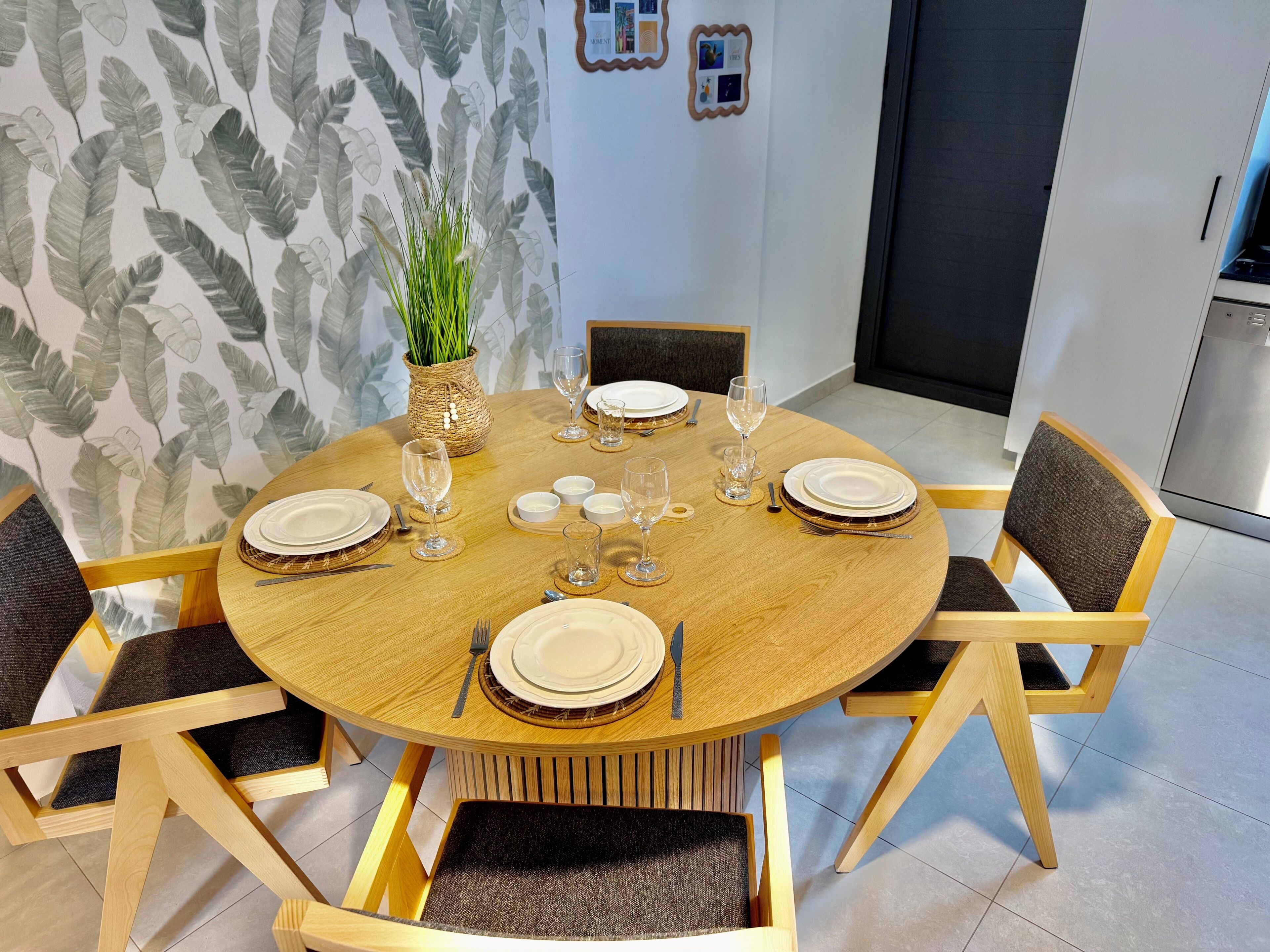 Een ronde houten eettafel, gedekt met borden, glazen en bestek, omringd door stoelen met donkere kussens.