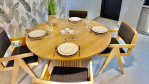 Uma mesa de jantar redonda de madeira posta com pratos, copos e talheres, rodeada por cadeiras com almofadas escuras.