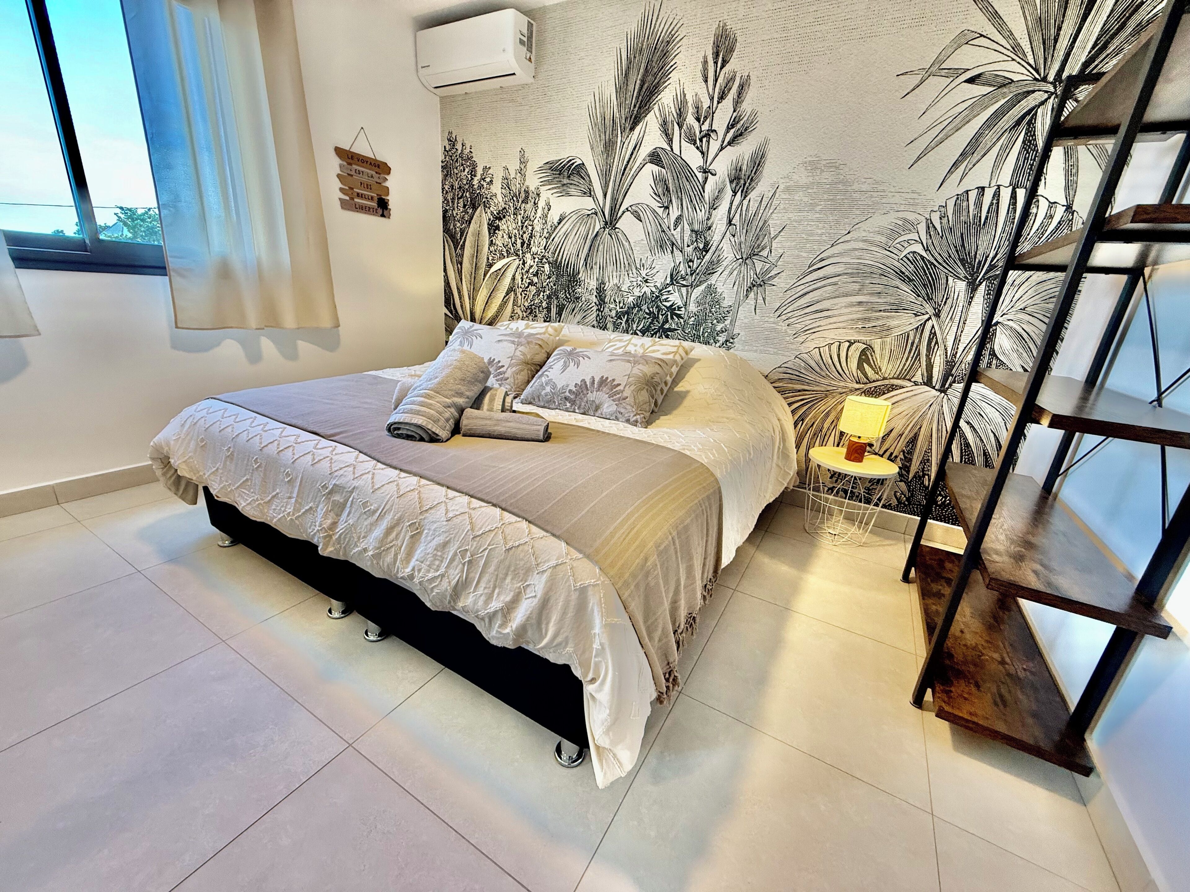 Un dormitorio con cama, mesita de noche, lámpara y un mural de plantas tropicales en la pared.