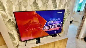 Uma televisão de tela plana sobre um suporte de madeira, com um controle remoto ao lado, em frente a uma parede com papel de parede estampado com folhas.