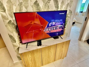 En platt-TV står på ett träställ, och en fjärrkontroll ligger bredvid. Bakom TV:n finns en vägg med tapet med ett lövmönster.