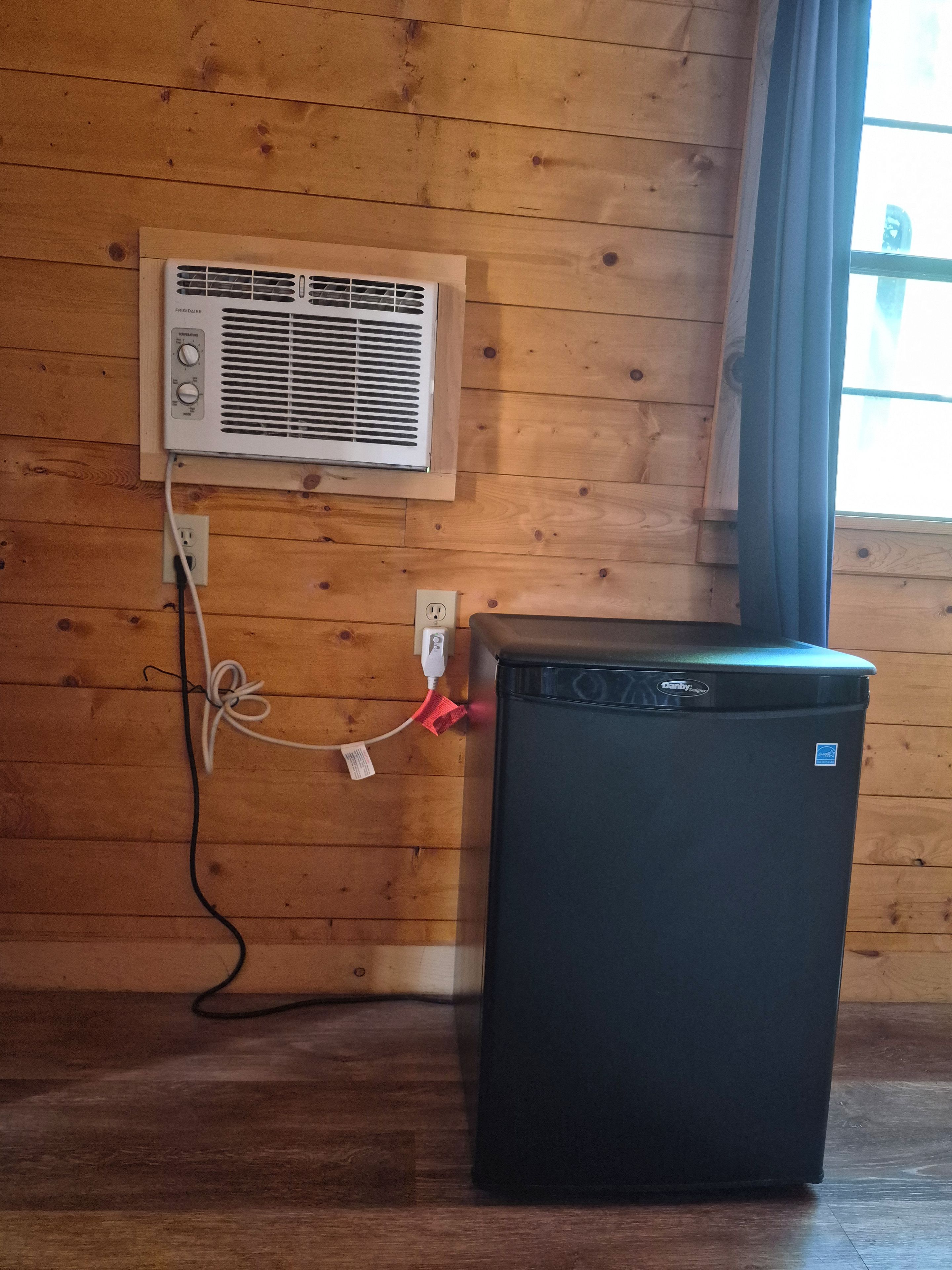Un refrigerador compacto con panel de control, conectado a un aire acondicionado de pared y un enchufe.