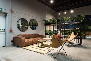Un espace de détente moderne avec un canapé en cuir marron, une table basse ronde et deux chaises pliantes. Deux miroirs ronds sont accrochés au mur, et une étagère noire abrite des plantes.