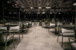 An indoor dining area with tables and chairs arranged in rows, large windows along one side, and a ceiling with recessed lighting. - Liberty (Nafplio)