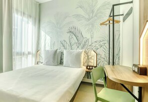 Quarto de hotel com cama, escrivaninha e cadeira. Uma parede apresenta um padrão de folhas tropicais.