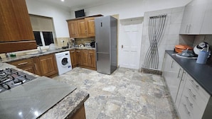 A kitchen with granite countertops, wooden cabinets, a stainless steel refrigerator, a washing machine, and a tiled floor. - Snoozeden Wellesley - Near to Slough Station (Cardiff)