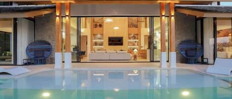 A modern house with a swimming pool, a glass-enclosed living area with a sofa and TV, and a stone facade.