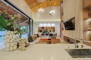 A modern kitchen with a built-in sink, a dining area with a sofa and chairs, and a large window overlooking a pool. - Chateau Du Village Patong (Krabi)