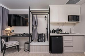 A compact hotel room with a four-poster bed, a desk with a lamp, a chair, a small trash bin, a compact kitchen with a microwave, a coffee maker, and a sink. - Chateau Du Village Patong (Krabi)