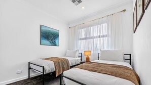 A hotel room with two beds, a bedside table with a lamp, a window with sheer curtains, and a wall-mounted painting. - The Art House | Courtyard + Alfresco Living (Campbelltown)