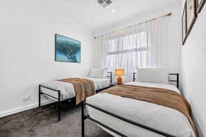 A hotel room with two beds, a bedside table with a lamp, a window with sheer curtains, and a wall-mounted painting. - The Art House | Courtyard + Alfresco Living (Campbelltown)