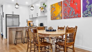 A modern kitchen with white cabinetry, a stainless steel refrigerator, and a wooden dining table with chairs. - The Art House | Courtyard + Alfresco Living (Campbelltown)