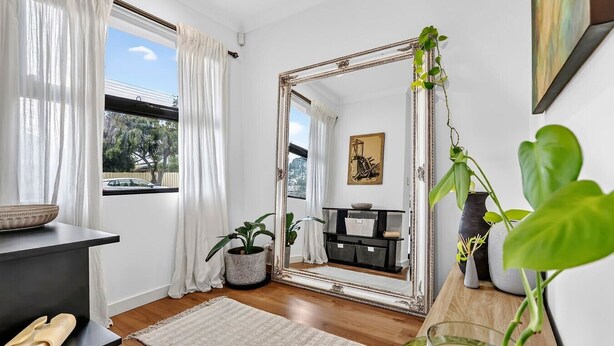 A room with a large mirror, a potted plant, a rug, and a window with curtains. - The Art House | Courtyard + Alfresco Living (Campbelltown)