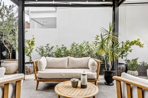 A modern outdoor seating area with a sofa, wooden chairs, and a round table. - The Art House | Courtyard + Alfresco Living (Campbelltown)