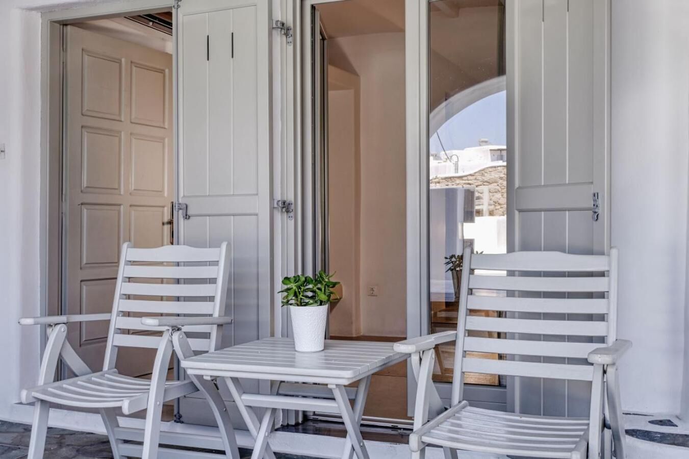 Un área de estar exterior blanca con dos sillas, una mesa, una planta en maceta y una puerta que da a un espacio interior.