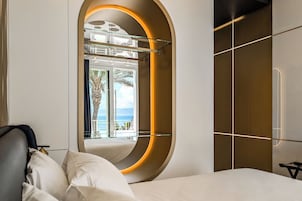 Habitación de hotel moderna con un espejo curvo, una cama con ropa blanca y vista al océano a través de grandes ventanales.