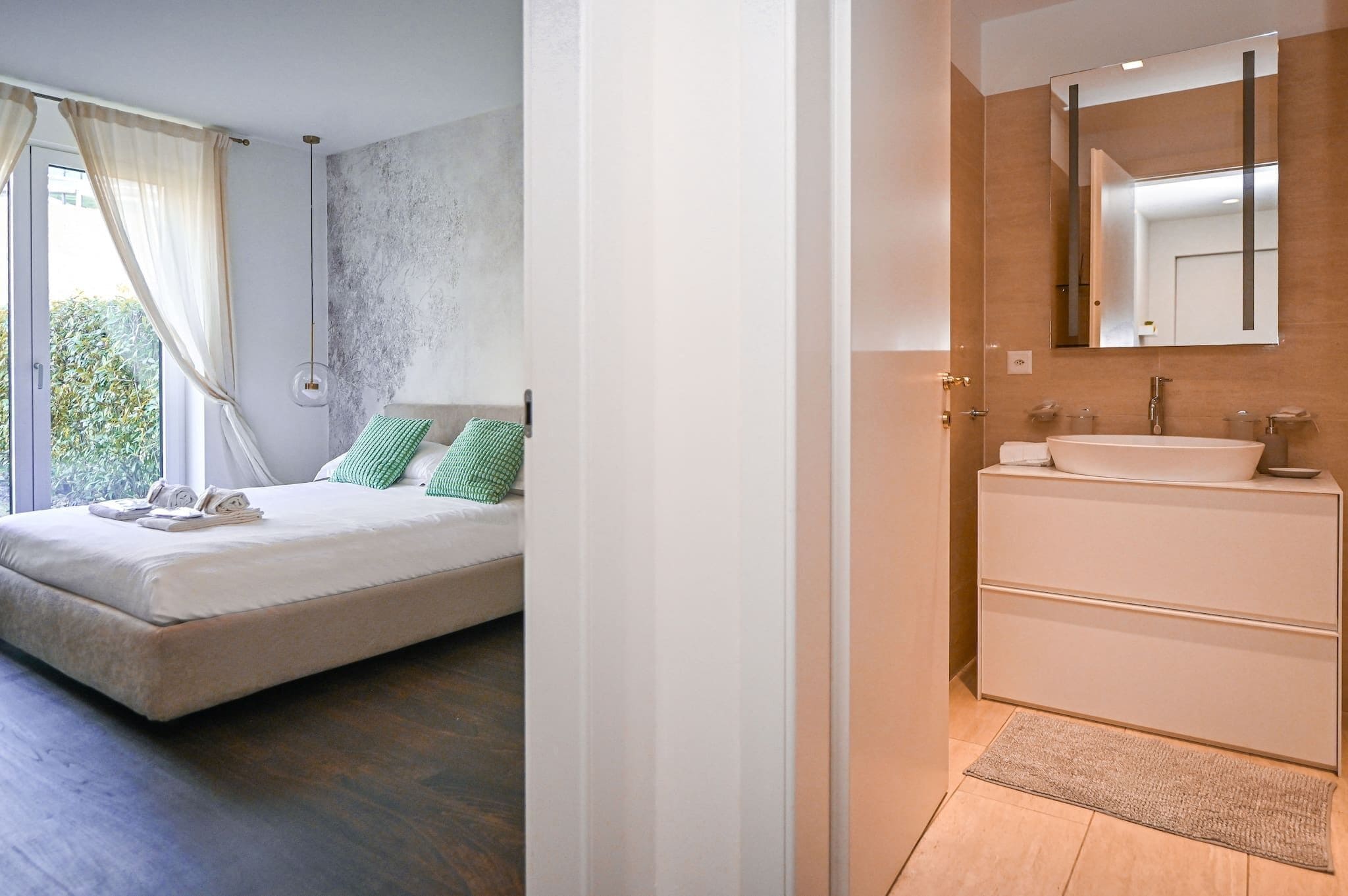 Ein modernes Schlafzimmer mit Bett, grünen Kissen und Blick ins Grüne. Das Badezimmer verfügt über einen großen Spiegel, ein Waschbecken und eine bodenebene Dusche.