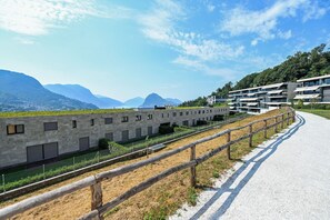 A modern building with a long, narrow structure, a paved pathway, and a fenced area with a view of mountains. - Luxury Corba Lake View Apartment With Terrace (Canobbio)