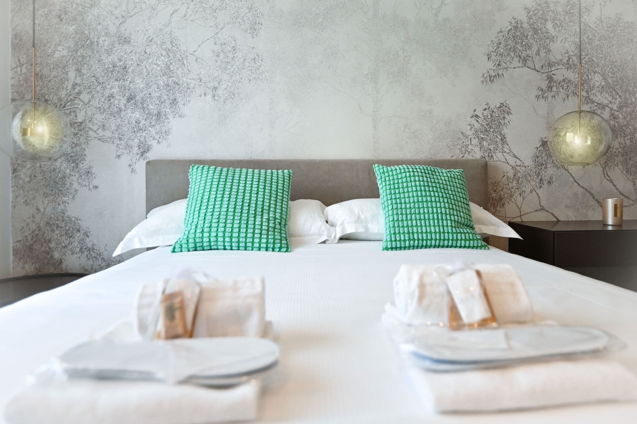 A hotel room with a large bed, two pillows, a nightstand, and a wall with a tree pattern.