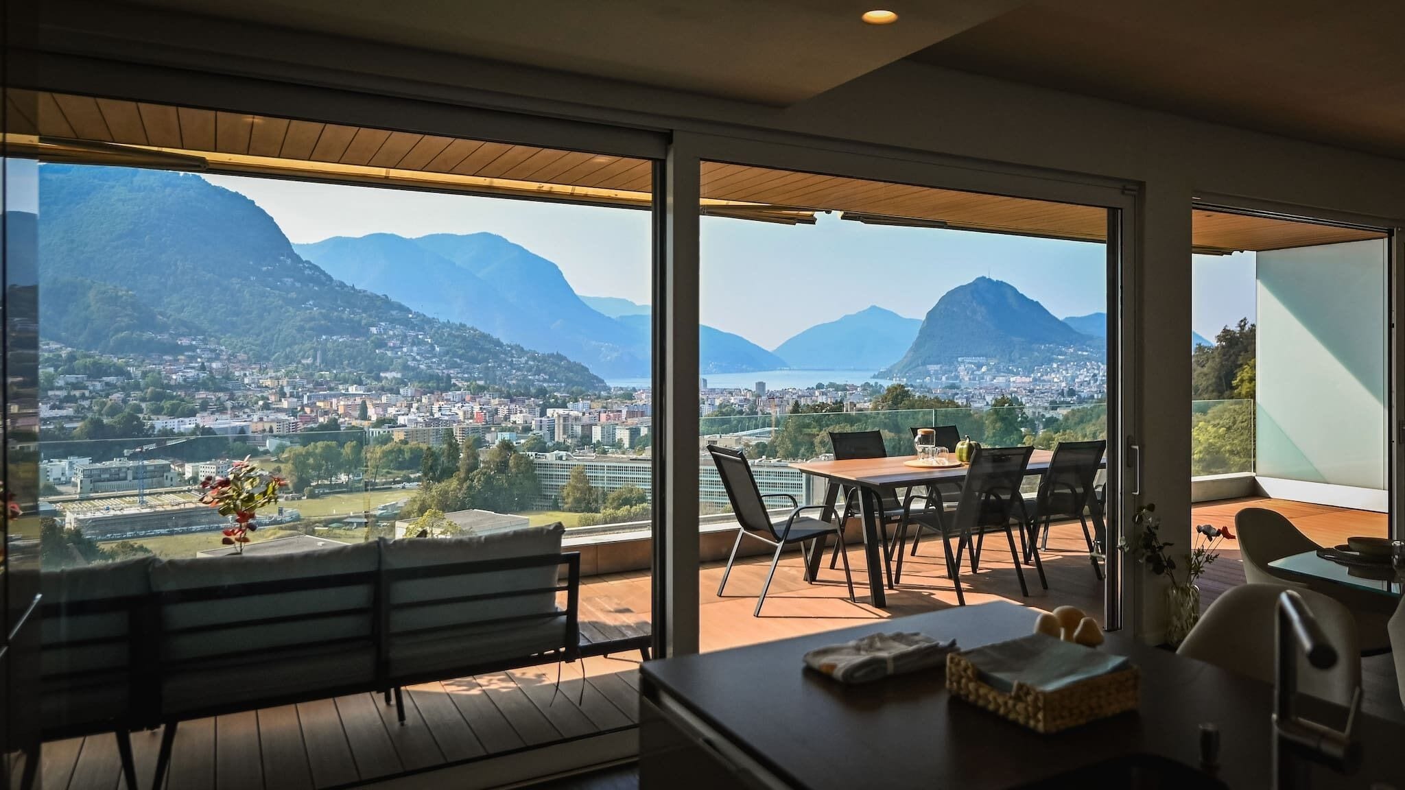 A balcony with a wooden bench, a table with chairs, and a view of a cityscape and mountains.