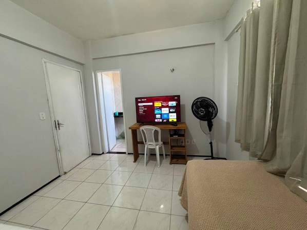 Una habitación con cama, un escritorio con computadora, una silla, un televisor y un ventilador.