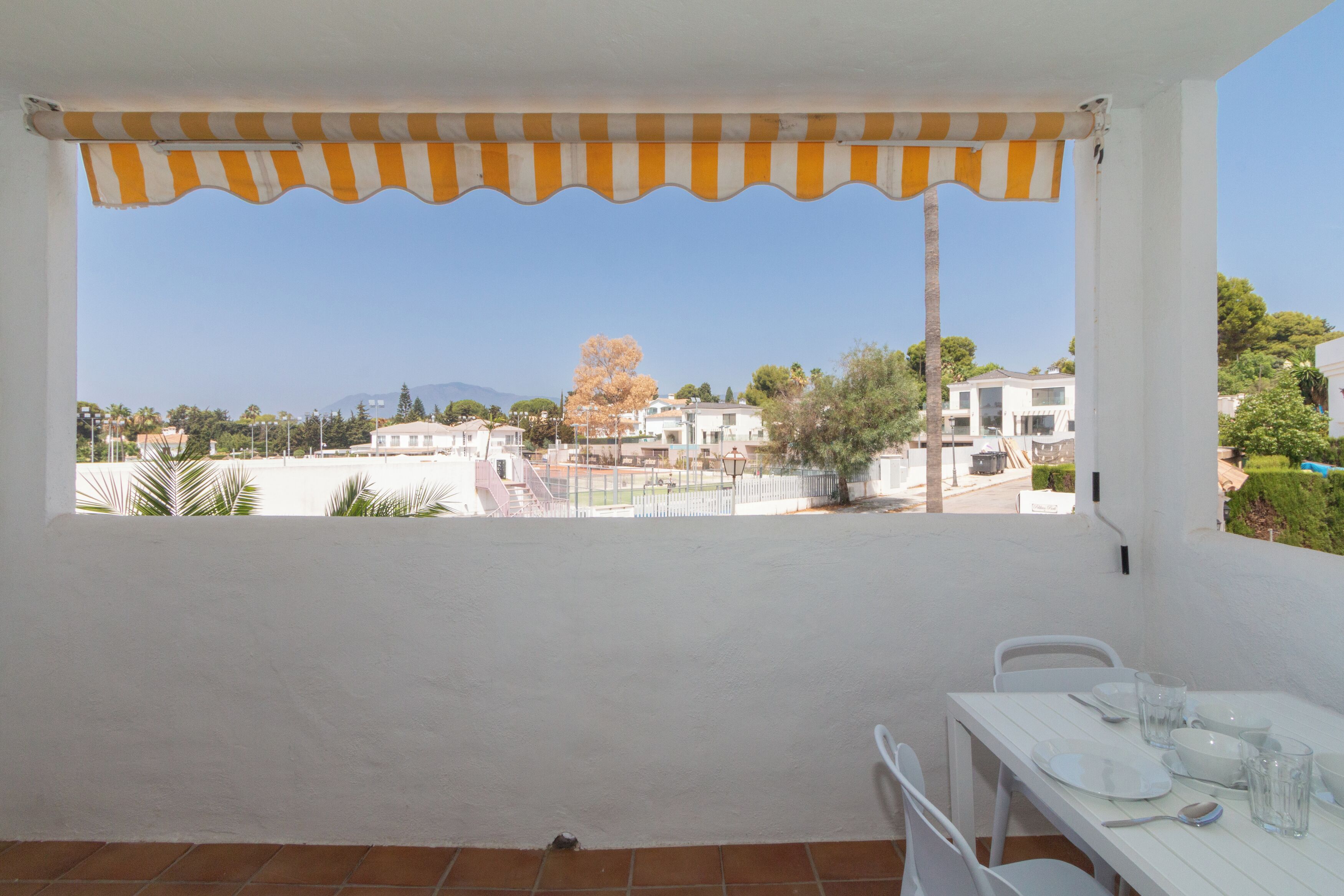 A balcony with a white table set for dining, overlooking a residential area with houses and a clear sky.
