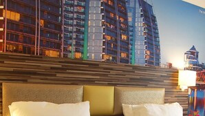 A hotel room with a large bed, a headboard with a textured design, and a view of a cityscape with tall buildings.