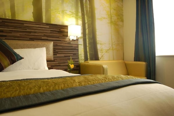 A hotel room with a large bed, a wooden headboard, a nightstand with a lamp, a sofa, and a wall with a forest mural. - Diamond Lodge (Manchester)