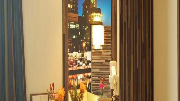 A hotel room with a desk, a chair, a vase with flowers, a mirror reflecting the cityscape, and a bedside table with a lamp.