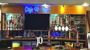 A bar area with a neon sign reading 'Cafe Bar,' a counter with a number '20' above it, and a variety of bottles and glasses on shelves.