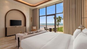 A hotel room with a large bed, a TV, a seating area, and a view of the beach.