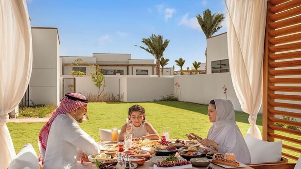Une famille est réunie autour d’une table pour déjeuner en plein air, avec une variété de plats et de boissons. L’ensemble est agrémenté d’un bâtiment moderne et d’une végétation luxuriante.