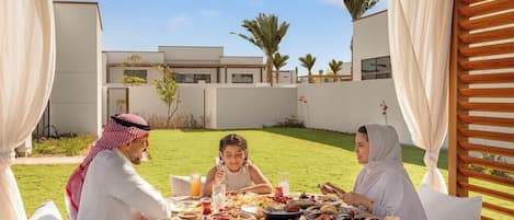 Una familia cena al aire libre con diversos alimentos y bebidas sobre una mesa, rodeada de un edificio moderno y vegetación.