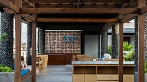 An outdoor seating area with a wooden pergola, stone pillars, and a reception desk with a sign.