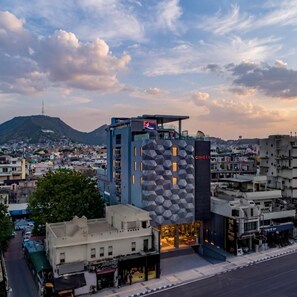 A modern hotel with a geometric facade, located in a cityscape with a mountain in the background. - Clarks Collection Grand Sapphire Ajmer (Ajmer)