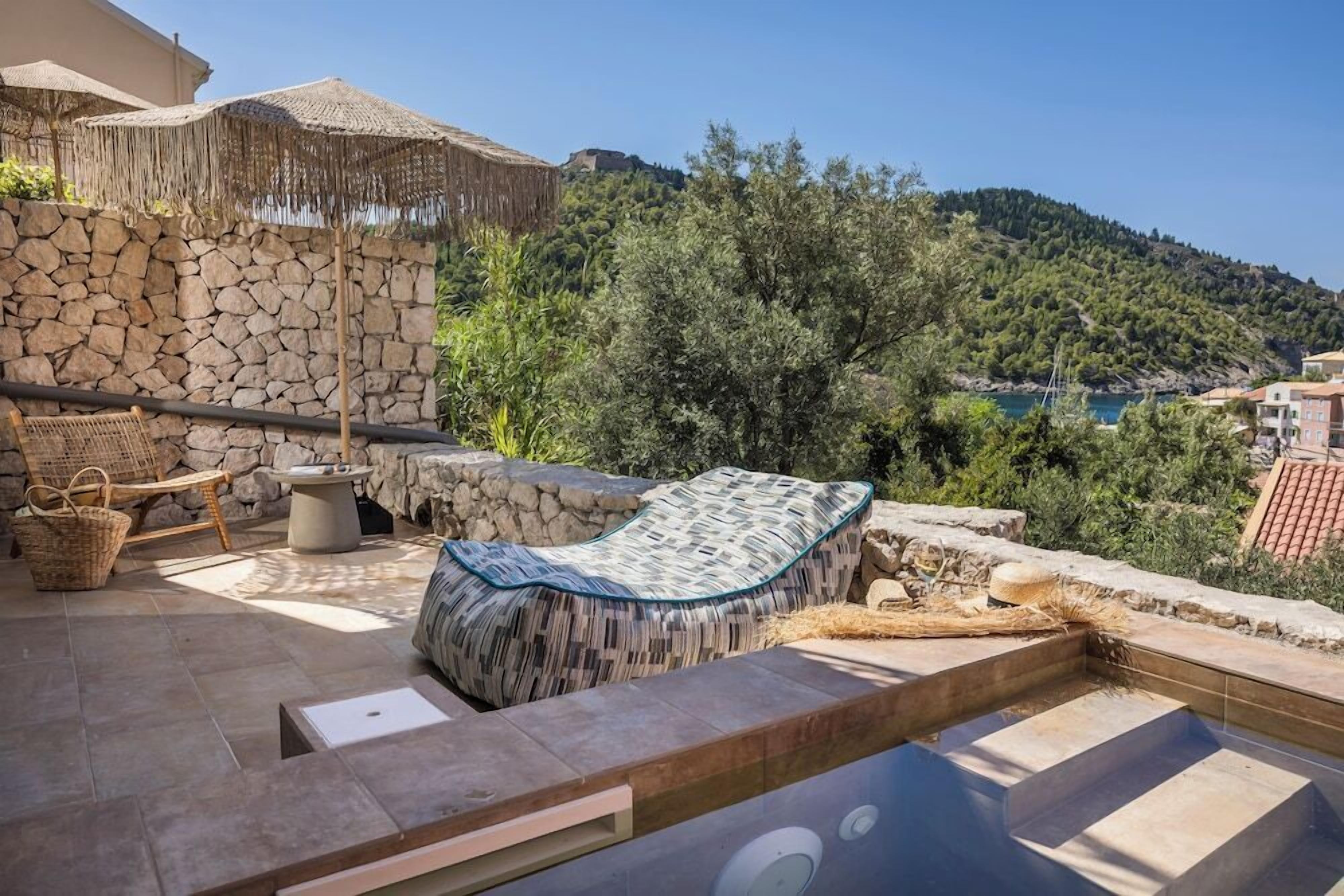 A stone wall, wicker furniture, a pool, and a view of a hill with trees and a body of water.