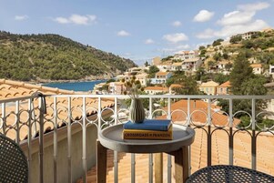 Un balcón con vistas a un pueblo costero, una mesa con un florero y libros, y una silla.