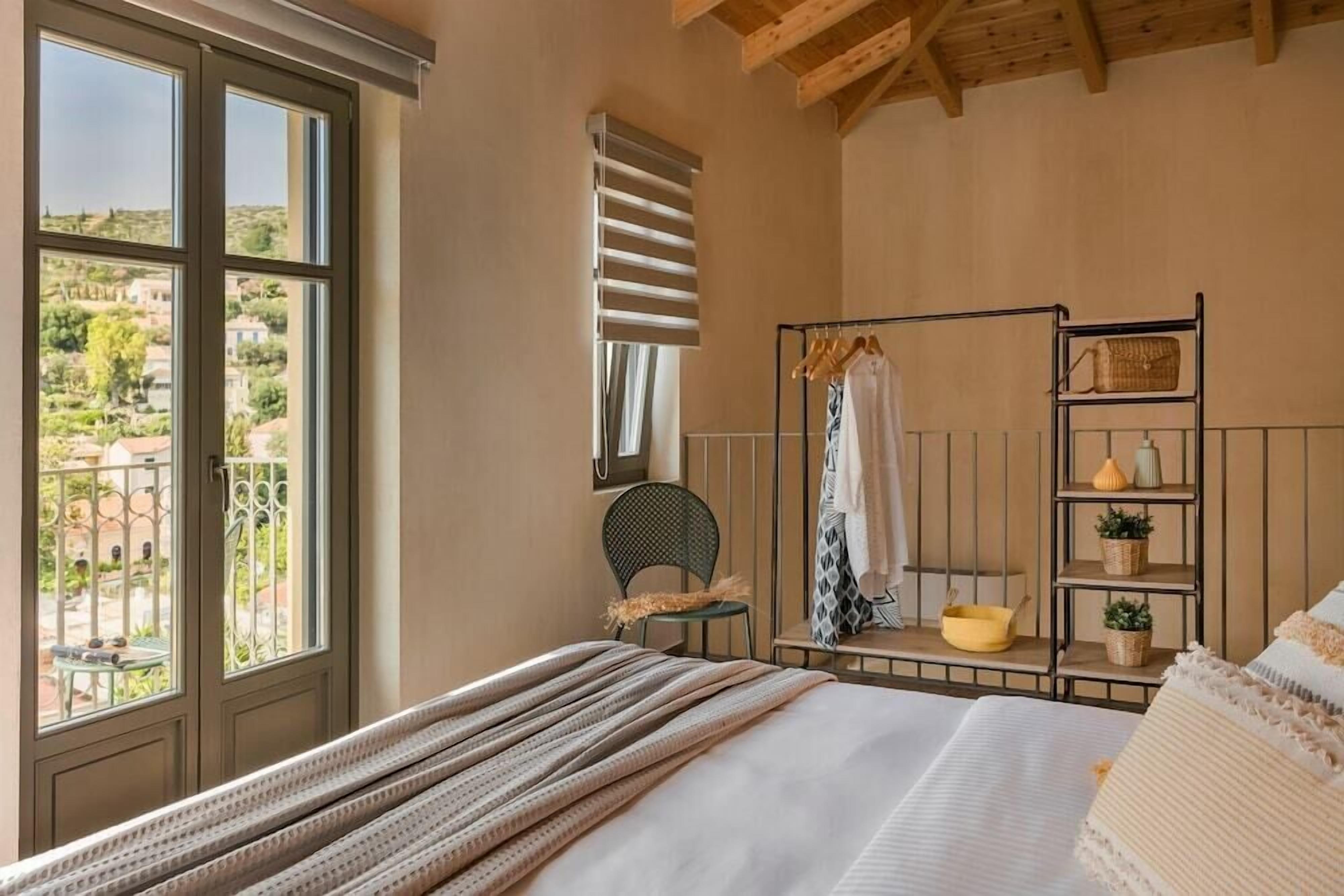 Un dormitorio con cama, ventana con vistas, una silla, estante con objetos y techo con vigas de madera.