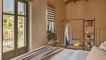 A bedroom with a bed, a window with a view, a chair, a shelf with items, and a ceiling with wooden beams.