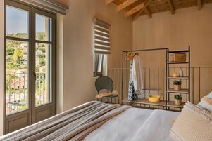 Quarto com cama, janela com vista, cadeira, prateleira com objetos e teto com vigas de madeira.