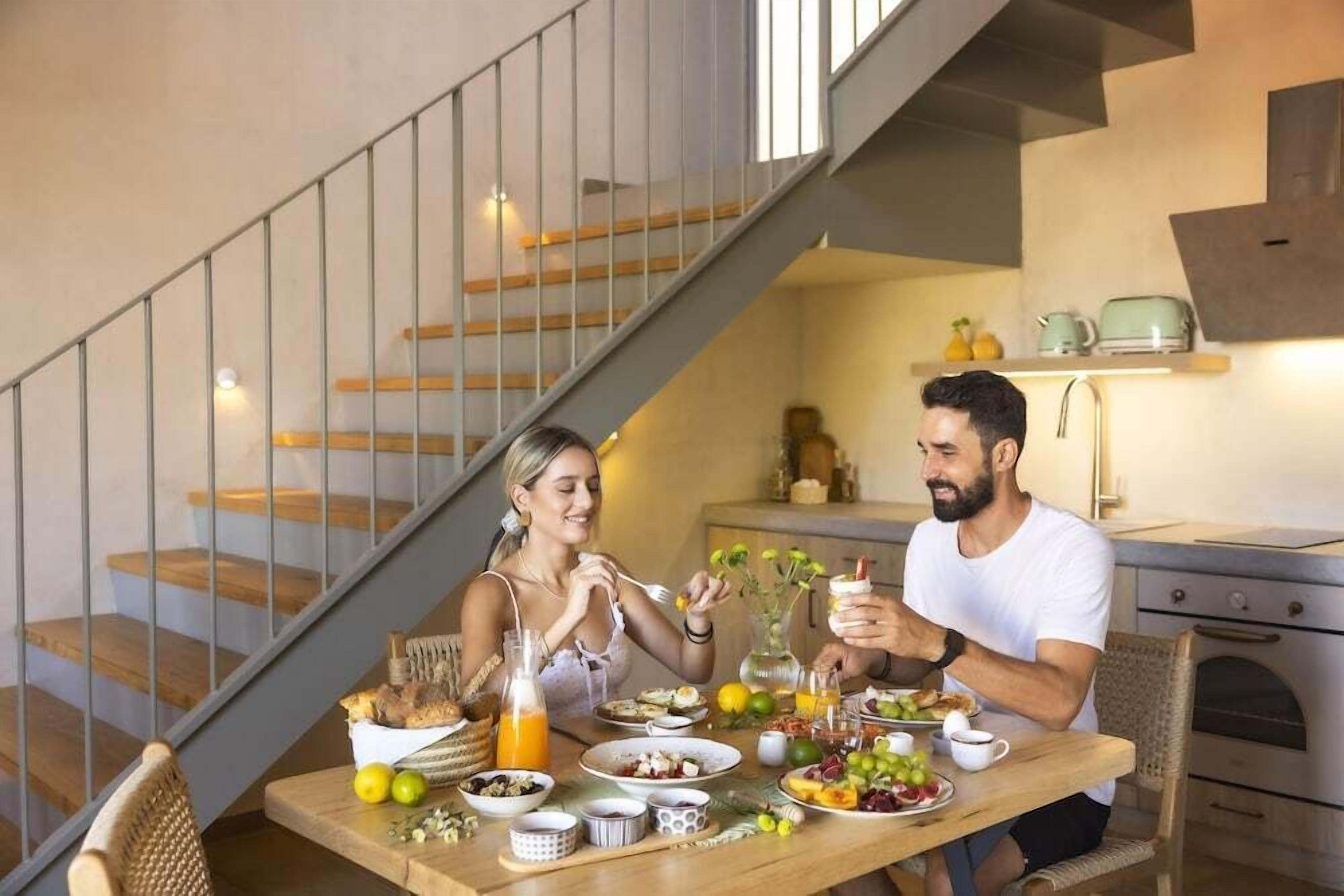 A dining area with a wooden table set for two, a staircase, and a kitchen in the background.